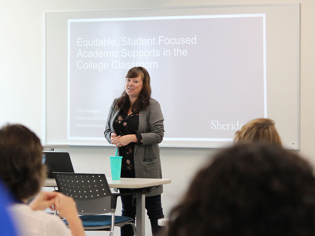 A person standing at the front of a classroom with a PowerPoint slide projected on the screen behind them.