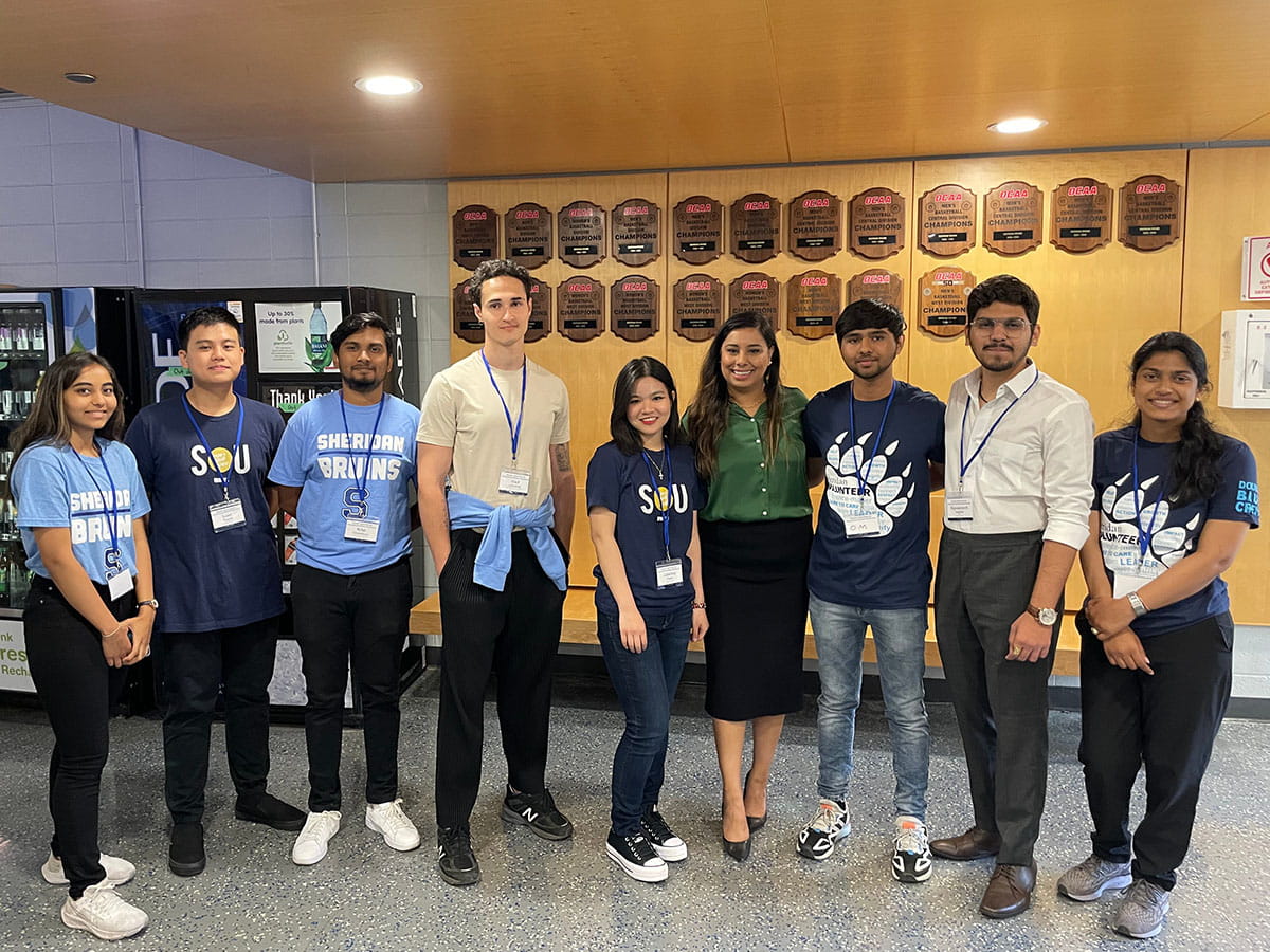 A group of students and MP Sahota posing for a photo in front of a series of plaques on the wall.