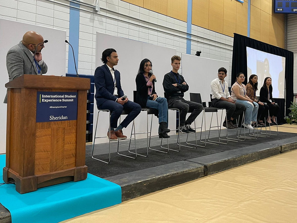 A panel of students seated side-by-side across a stage. The moderator stands to the left behind a podium.