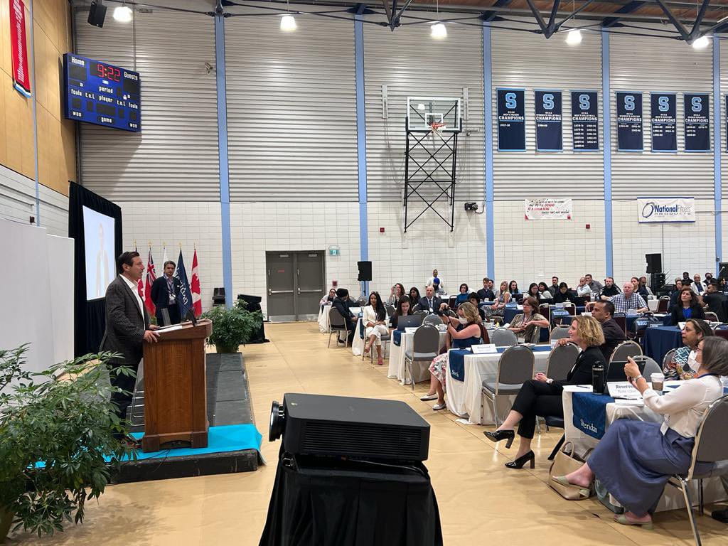 Patrick Brown stands to the left speaking behind a podium. Spectators are seated at tables on the right.