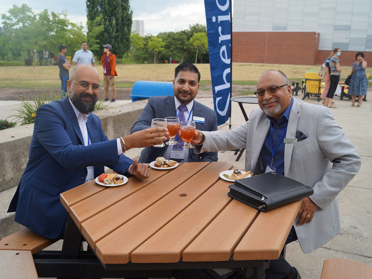 Three people seated at a picnic table outside raising their glasses in a toast.
