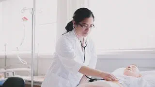 A student nurse using her stethoscope to examine a patient-care mannequin in a bed