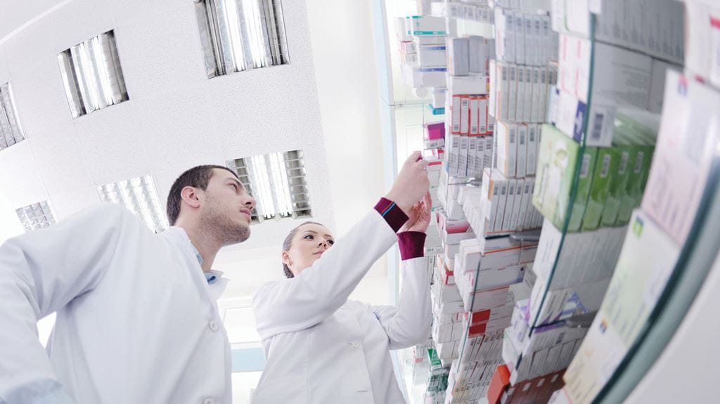 Two pharmacy students in lab coats looking at shelves of pharmaceutical supplies