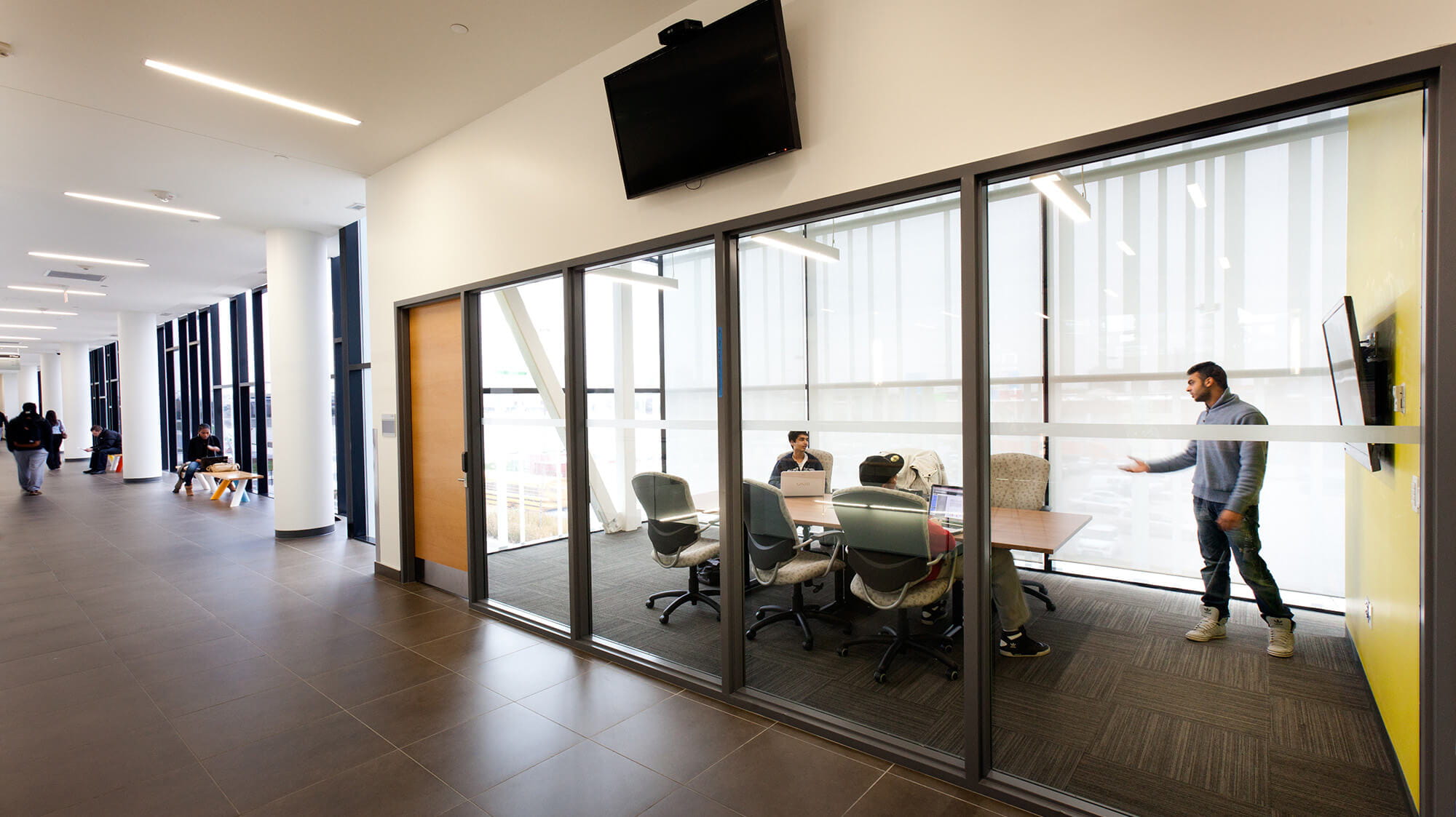 A hallway looking into a glass-walled group study room with a table and students inside.