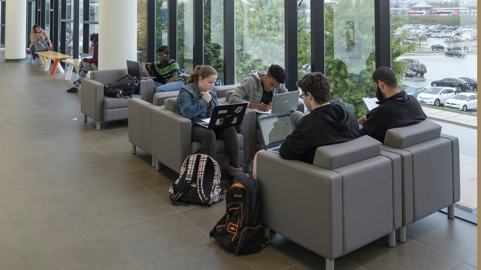 Students with latops sitting in comfortable chairs in a hallway with tall bright windows