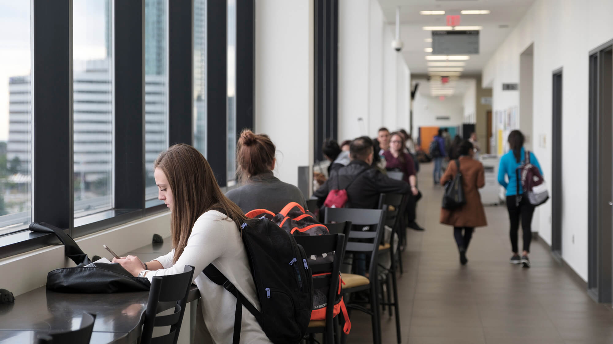 Students sitting at tables along the hallway beside bright tall windows