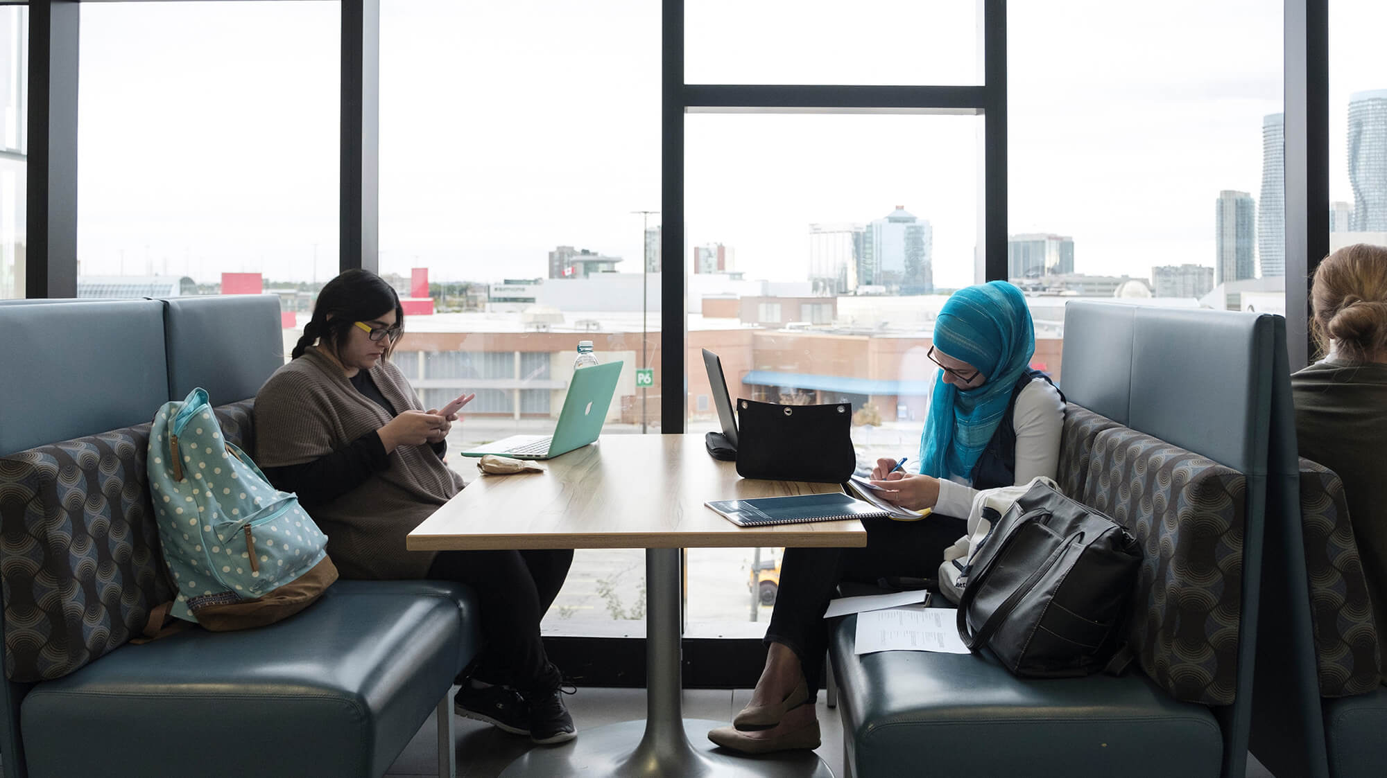 Two students sitting across from each other and studying in a booth with bright tall windows