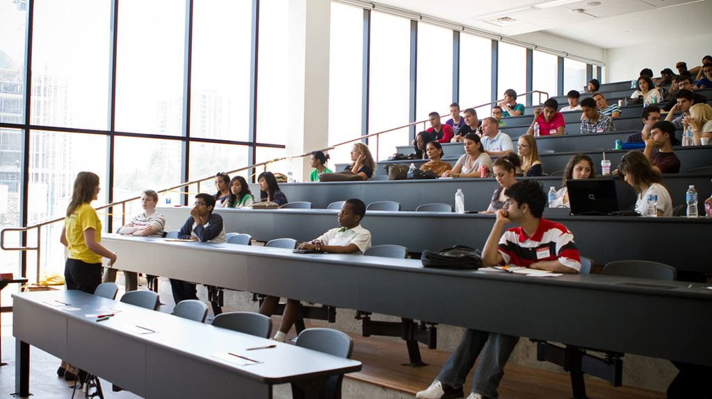 Many students and a teacher in a tiered lecture hall with a wall of windows