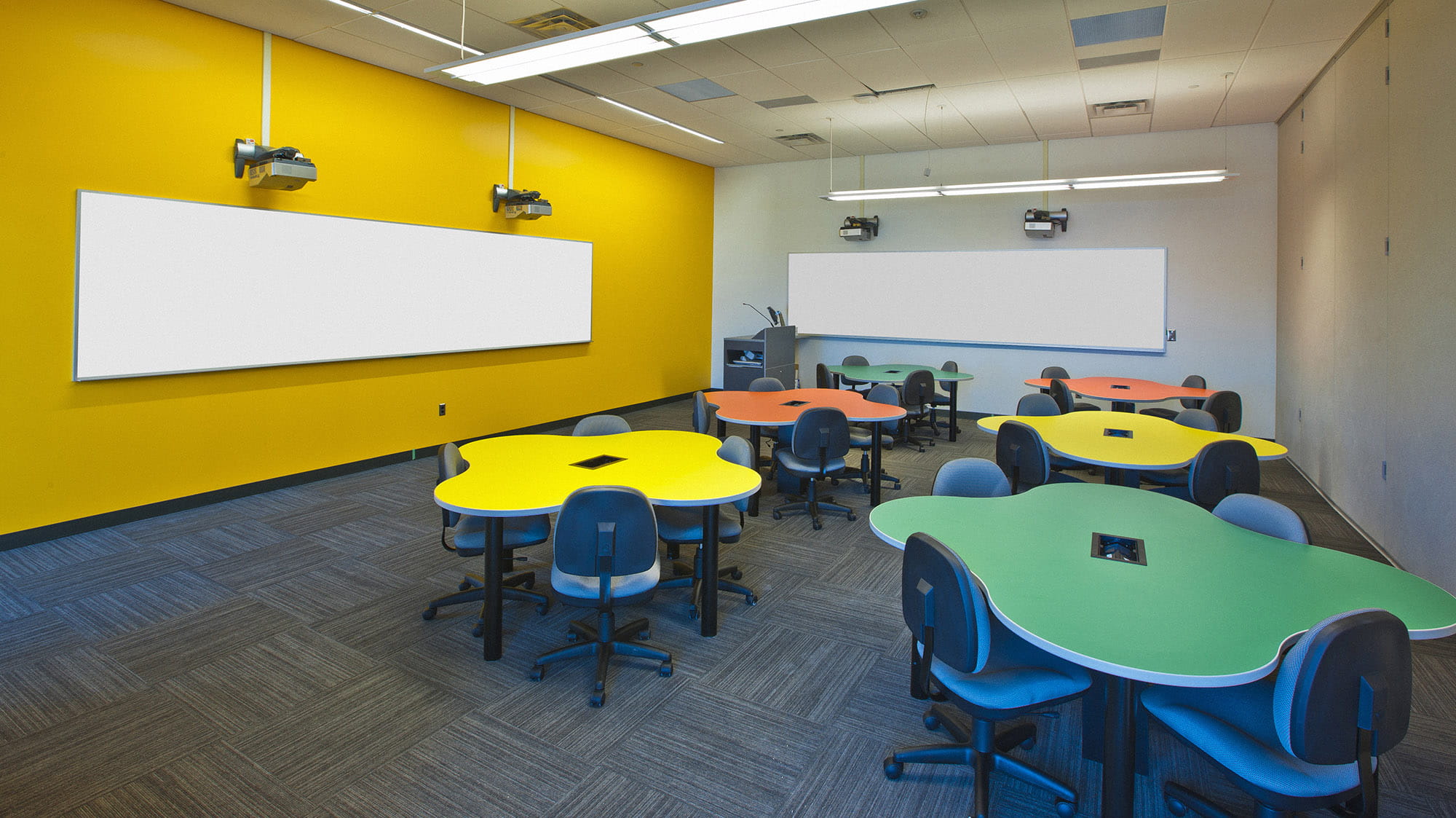 A classroom with a yellow wall, filled with multicoloured amoeba-shaped tables.