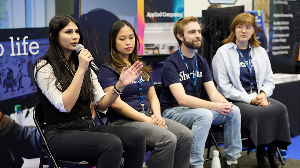 Four people sitting side-by-side – one is speaking into a microphone.