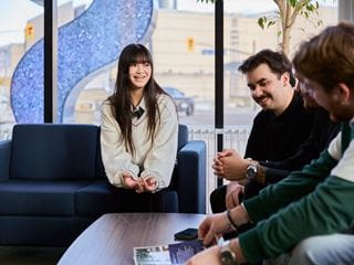 Four students sitting and talking on couches with a large Sheridan blue 'S' visible through the window behind them.