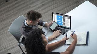 Two students working side-by-side at a desk with a laptop and a notebook.