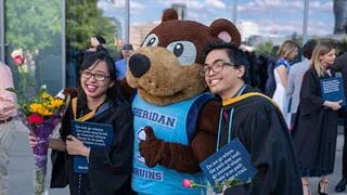 Two Sheridan grads in cap and gown posing for a picture with Bruno, Sheridan's mascot
