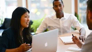 Three people in conversation. The person on the left has a laptop open on the desk in front of them.