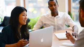 Three people in conversation. The person on the left has a laptop open on the desk in front of them.