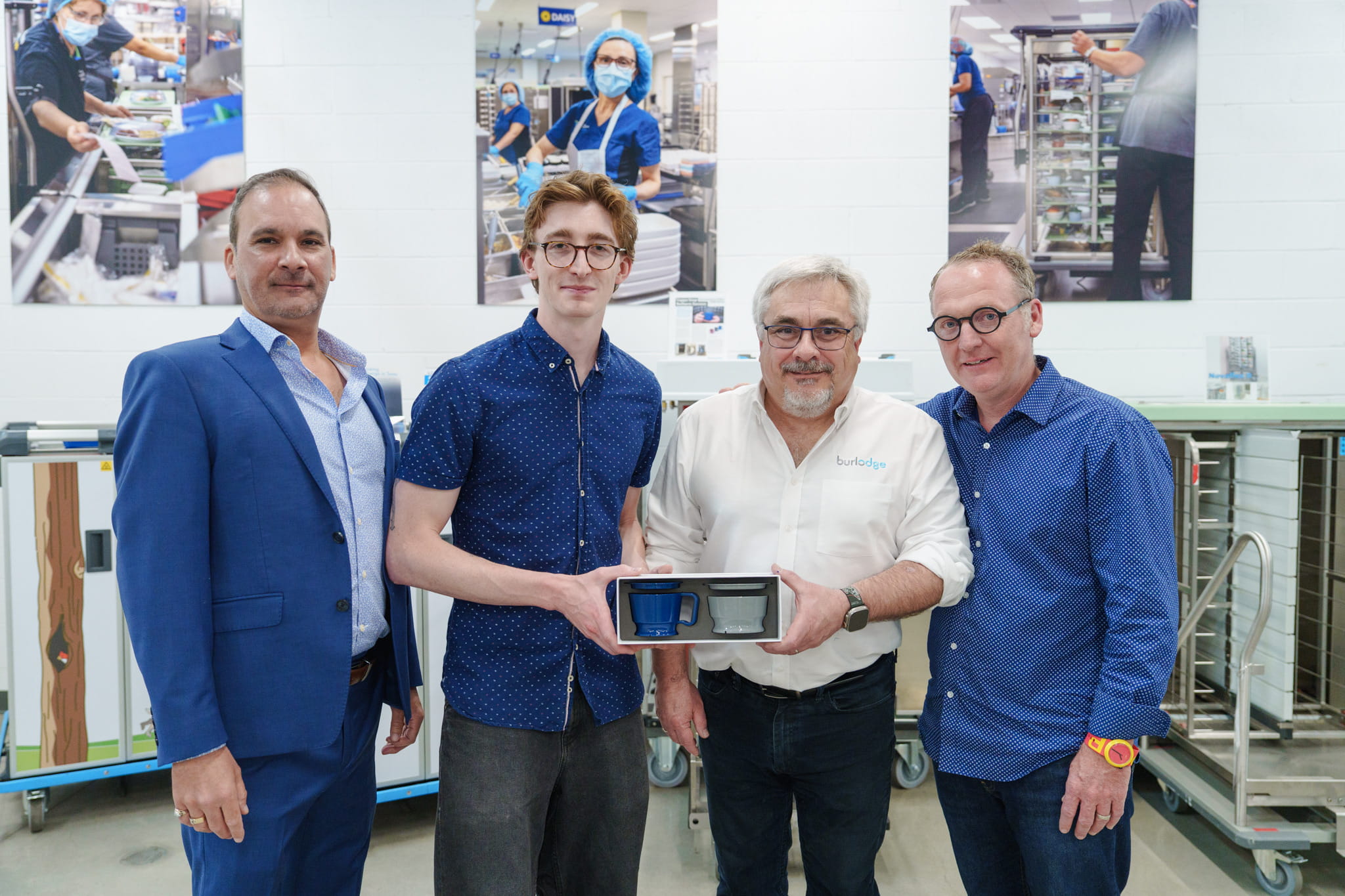 A group photo picturing Neil Sapra, Tobin Nickle-Lott, Paul Gauntley and Scott Currie holding samples of a new cup and bowl system with other Burlodge equipment and branding in the background