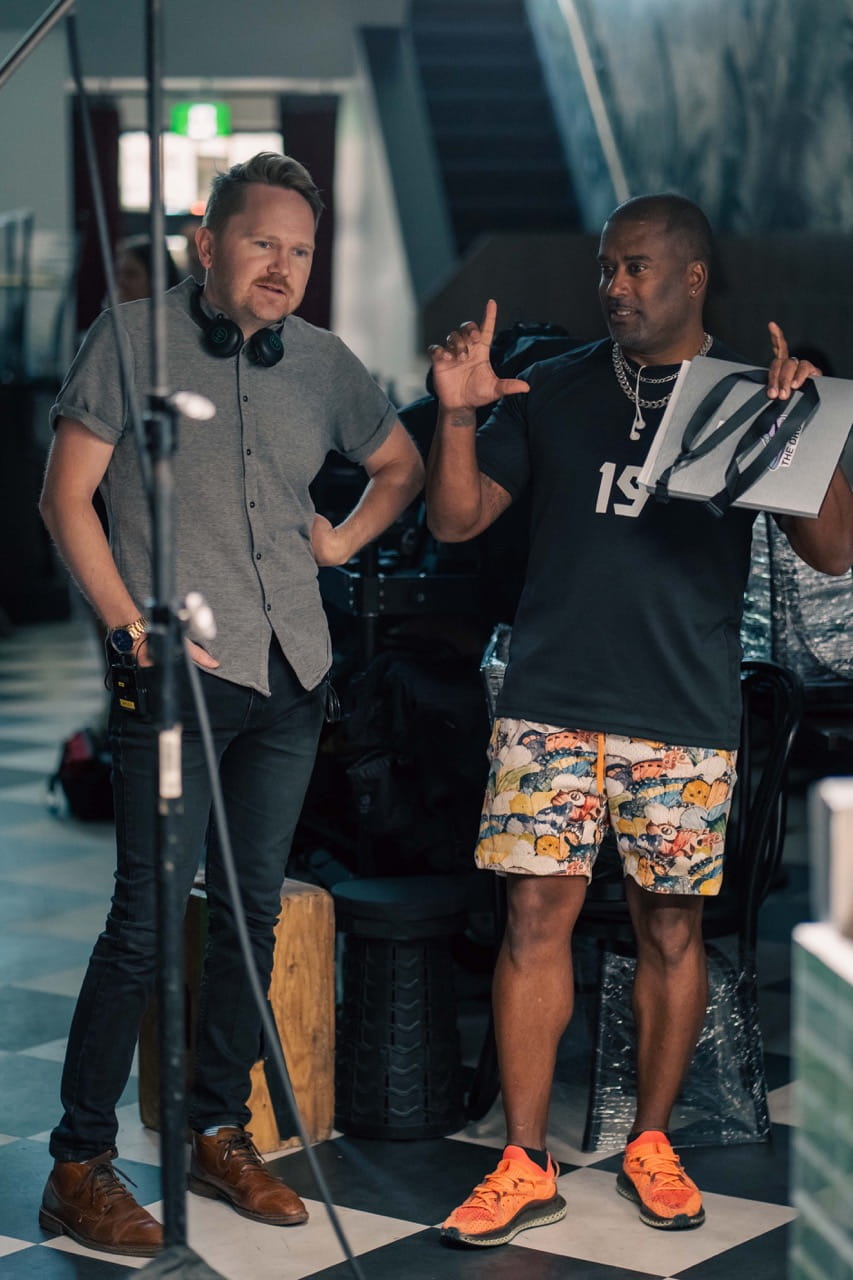 David Mewa, dressed in a t-shirt and shorts, interacts with a crew member on set of The Millwood Murders: Buried Truth