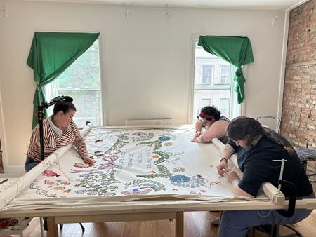 Three artists bend over a table as they work on a large embroidery project.