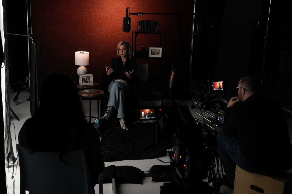 Dr. Samantha Nutt sits in a chair in front of a red wall in a dimly-lit room while being interviewed by Laila Vaga