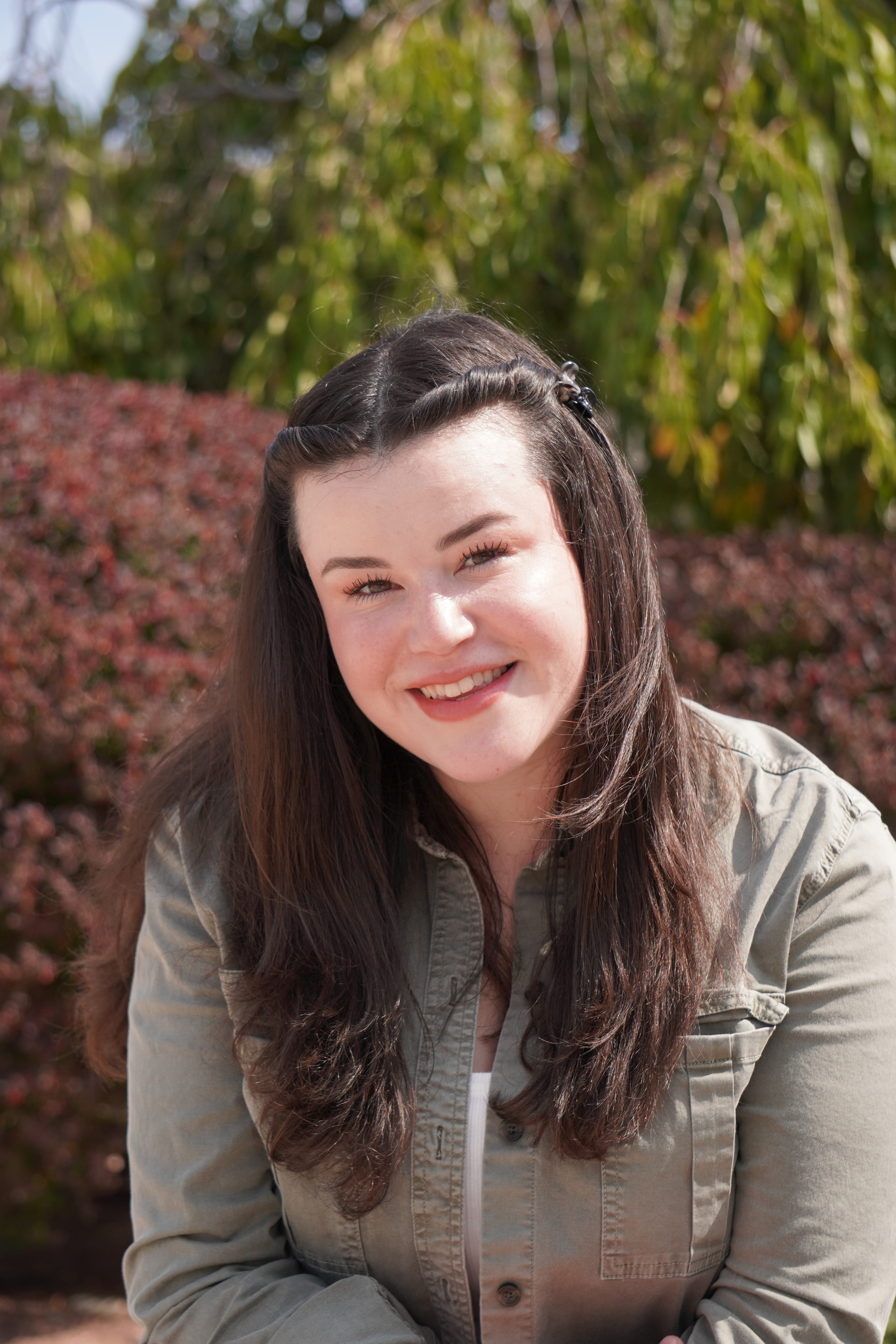 Sydney Brown sits on a bench in front of a tree and bush at Sheridan's Trafalgar campus.