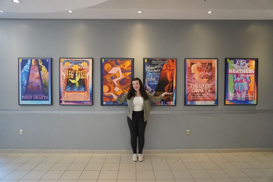 Sydney Brown stands in the middle of six posters on a wall showcasing theatre shows happening at Sheridan.