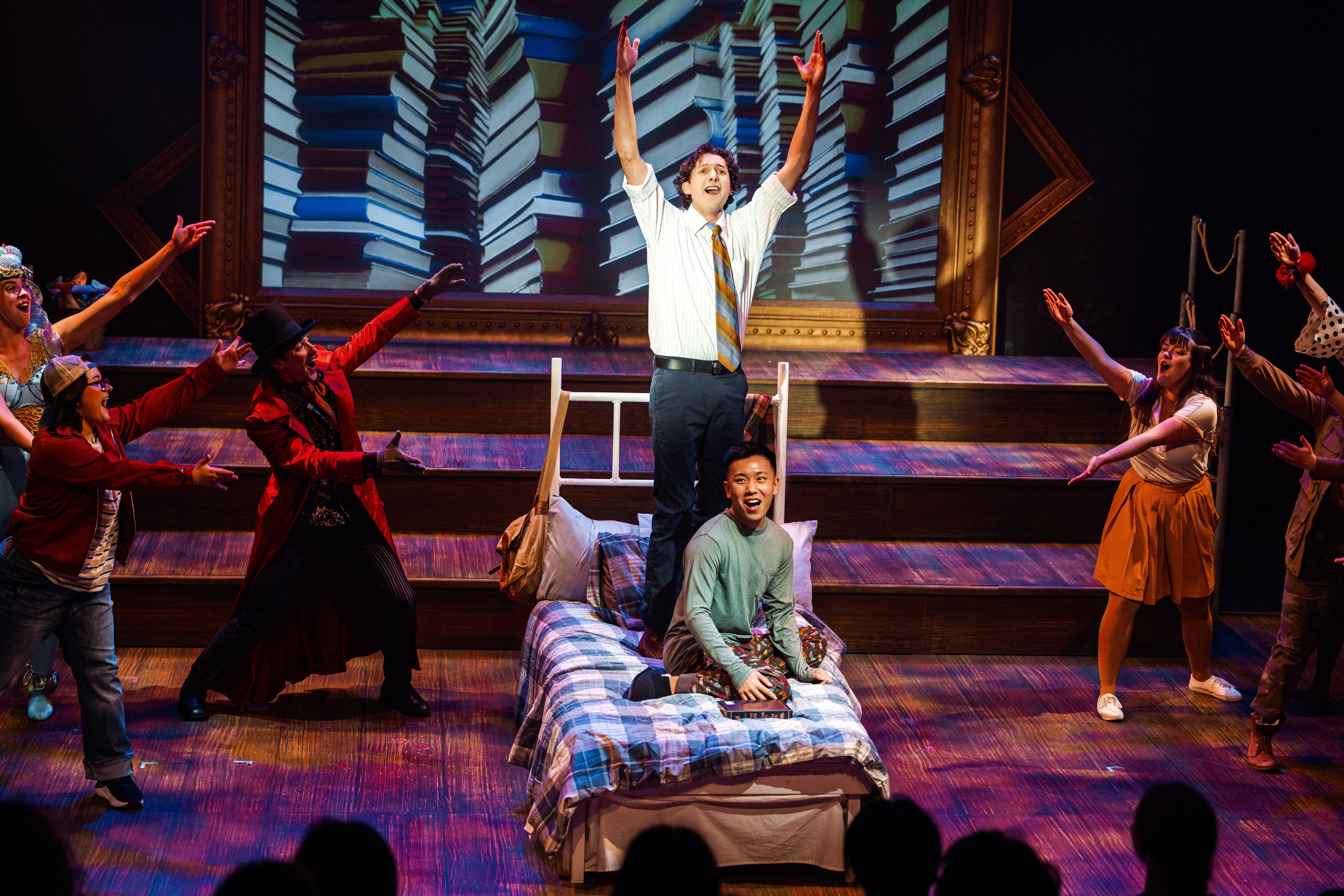 An actor stands on a bed on stage and another actor sits on the bed. Both are singing.