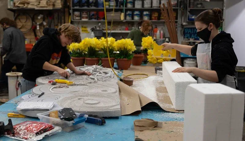 Three students work at a table preparing props for production. There are several pots with yellow flowers on the table.