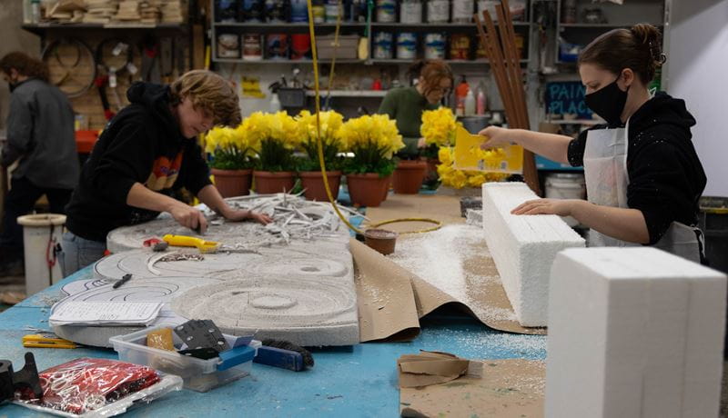 Three students work at a table preparing props for production. There are several pots with yellow flowers on the table.