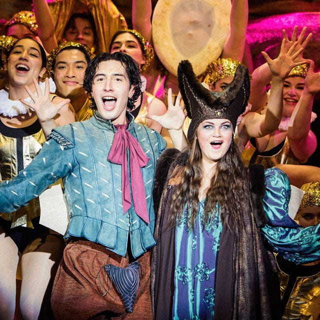 Two actors from the production of Something Rotten! are surrounded by chorus members as they sing.