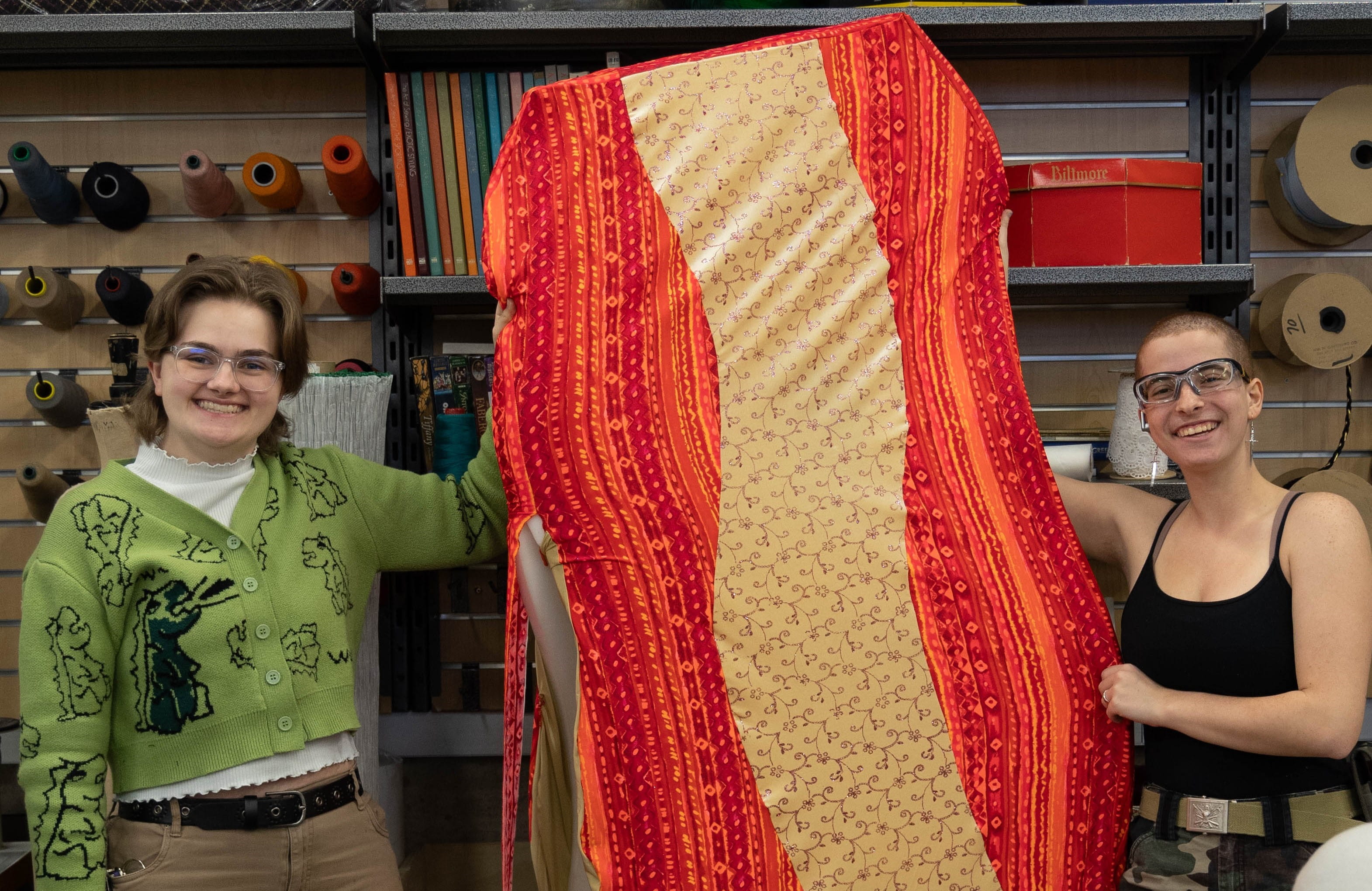 Two people hold a large costume made of fabric that looks like a slice of bacon.