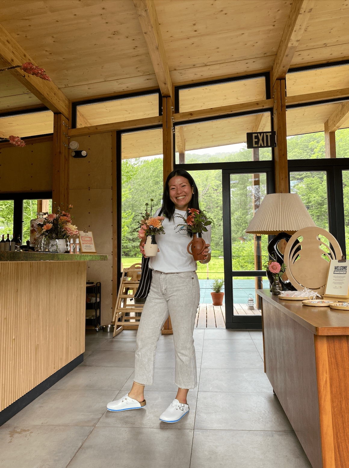 Christine Lui holds two vases containing flowers while standing inside an office area with large windows in the background
