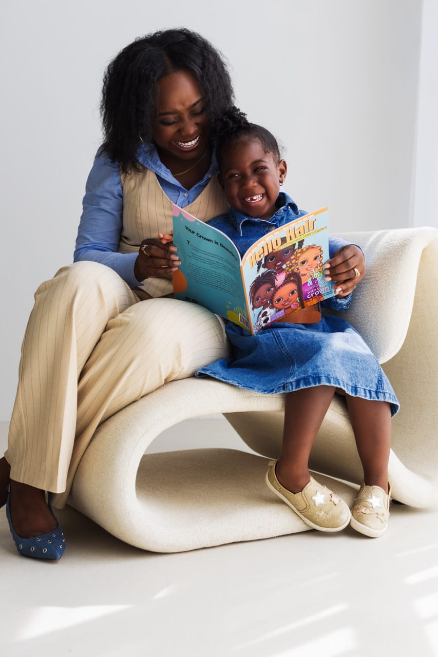 A child looks up and smiles as she reads Hello Hair while sitting in a chair beside Anita Grant