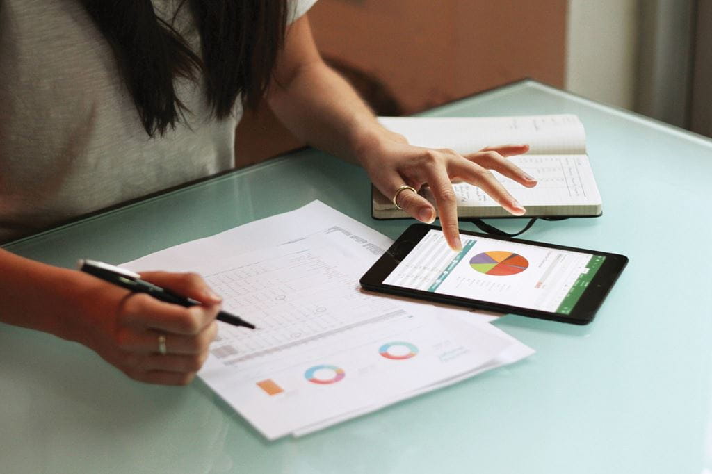 A glass desk with a notebook, printed documents and tablet on top. A person sitting at the desk is reviewing the various charts and data on the printed documents and tablet.