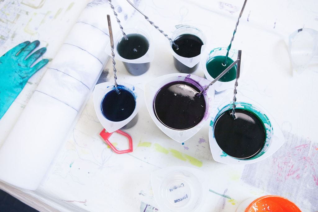 Green, purple and blue paint in containers on a table, with a paint stained rubber glove placed next to it