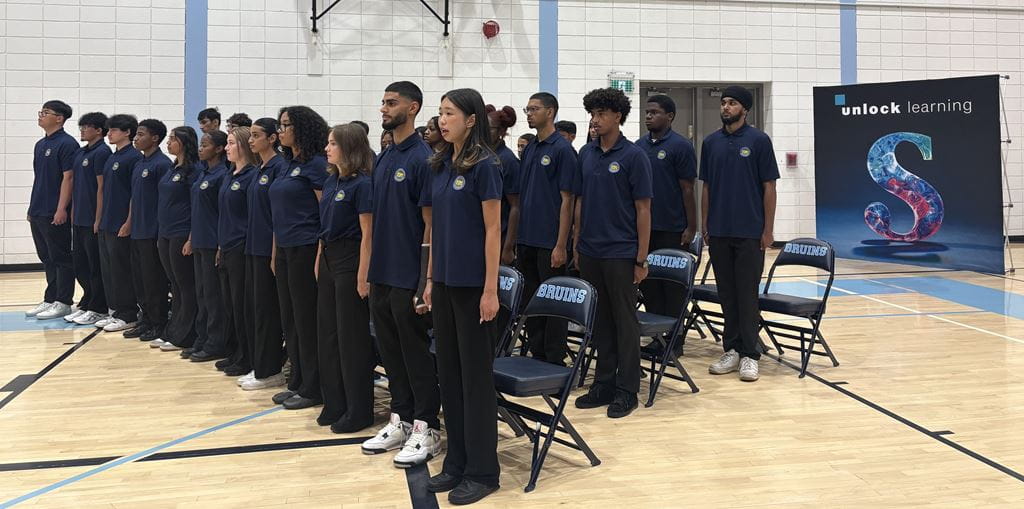 A group of 35 students wearing navy stand in three rows in front of their chairs in a gym.