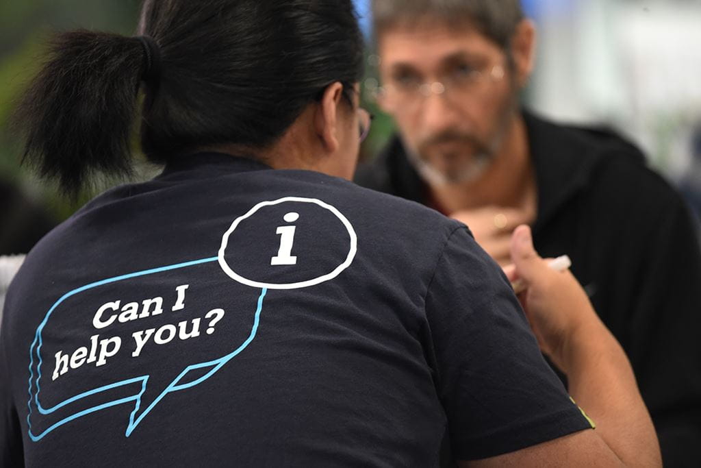 A Sheridan employee wearing a dark blue t-shirt that says "Can I help you?" while assisting a Fall Open House attendee