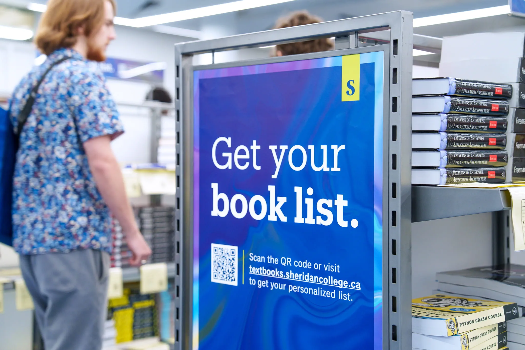 Students walk past a shelf filled with textbooks that has a large blue and purple sign that states "Get your book list" in bold white font.