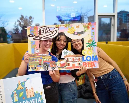Three students smile while looking through a cutout in a decorative poster that represents Colombia