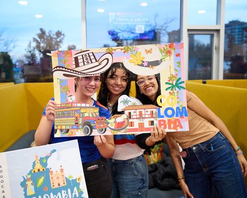 Three students smile while looking through a cutout in a decorative poster that represents Colombia