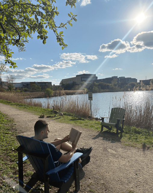 Joshua Colliopoulos is pictured seated in a Muskoka chair reading a book beside the pond at Sheridan's Davis Campus
