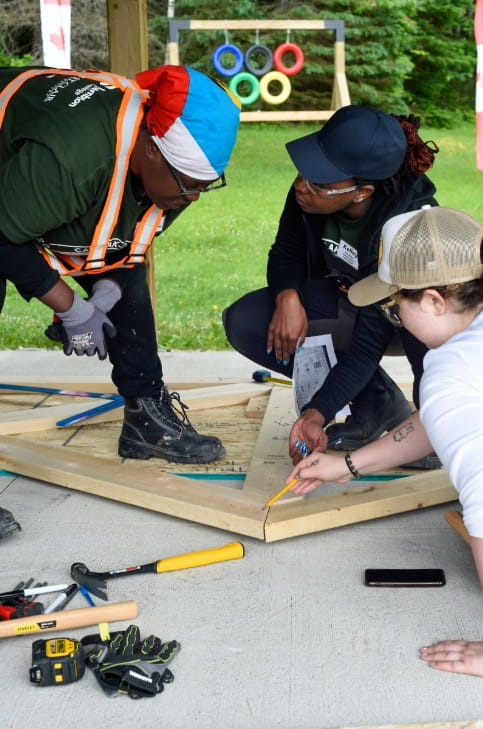 Three people kneel down while assembling a roofing truss.