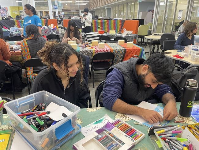 Two students sit at a table with art supplies. One student watches as another creates an image on paper.