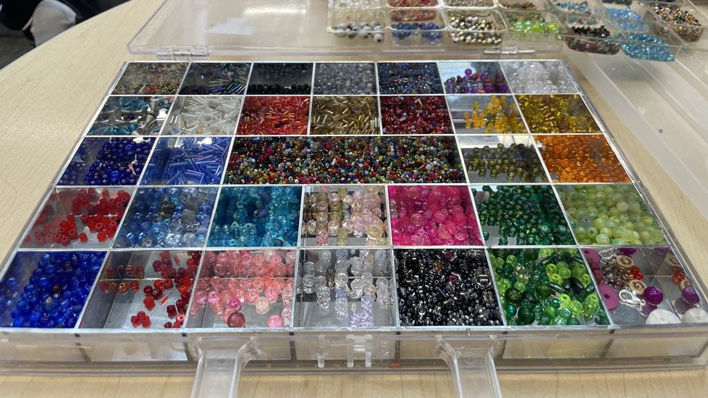 A square container with many small square slots containing colourful beads.