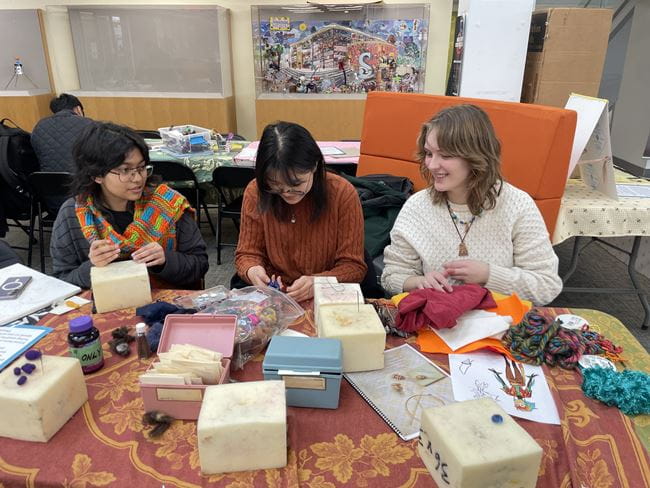 Three students sit at a table covered in art supplies while smiling and working on a project.
