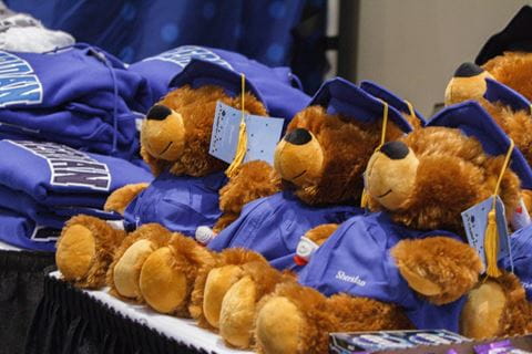 A close up image of stuffed teddy bears wearing graduation caps and gowns, next to Sheridan logo hoodies.