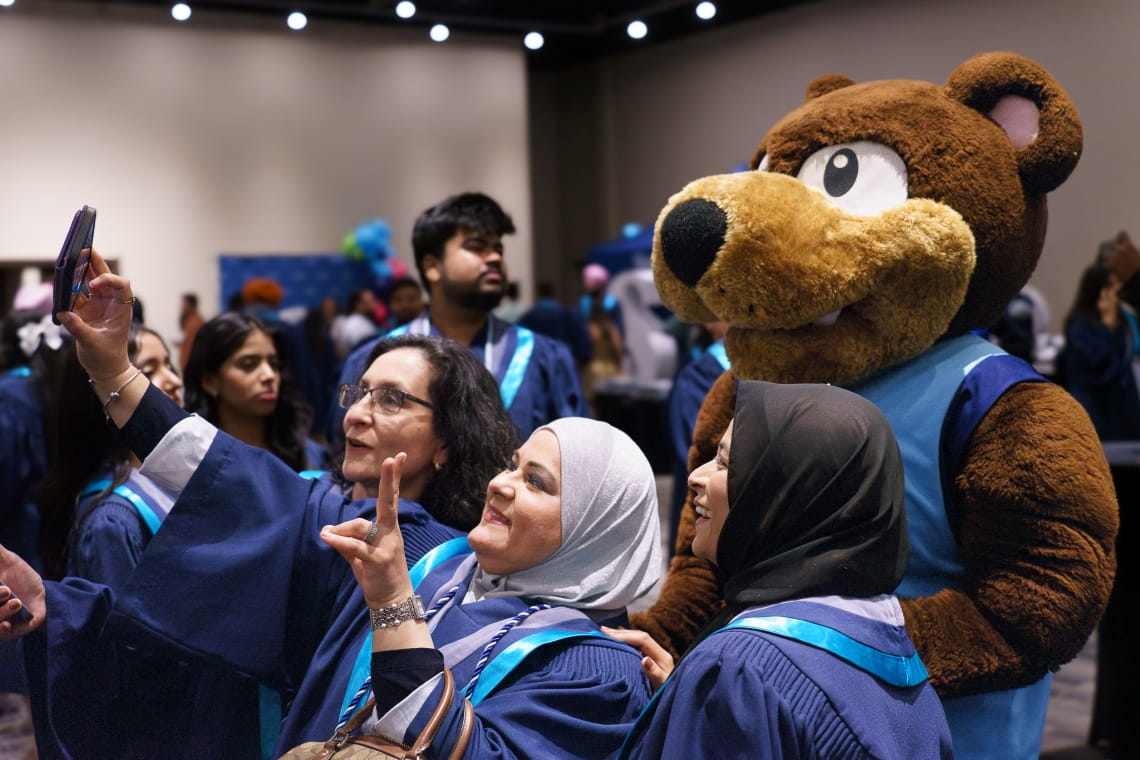 Three ladies wearing grad gowns pose and smile for a selfie with Bruno.