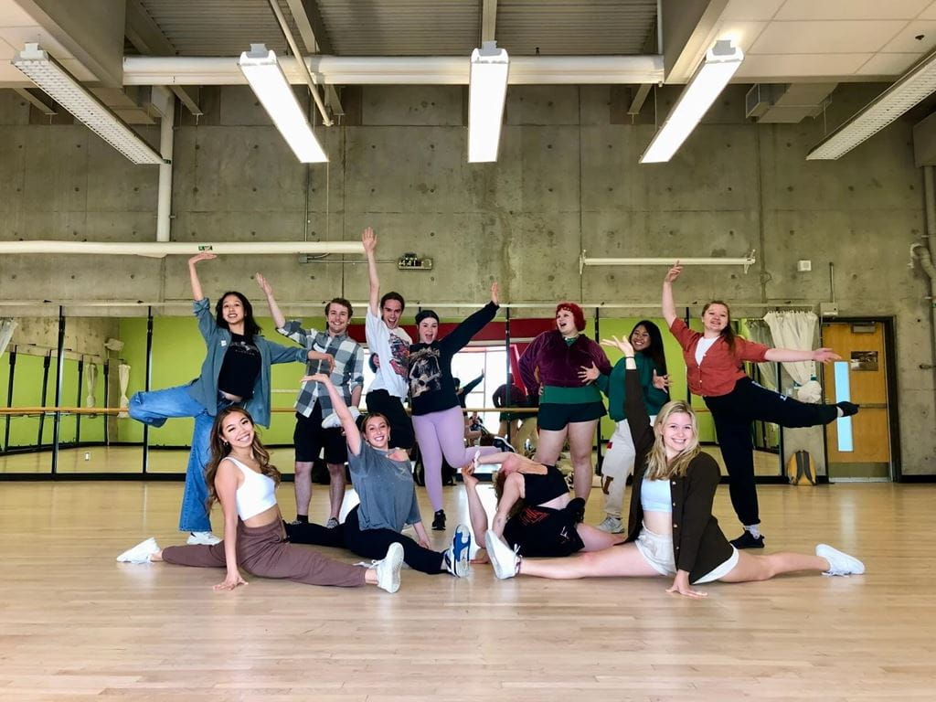 A group of theatre camp participants pose together in a dance studio with mirrored walls and a wood floor. Several participants stand with arms extended in performance-style poses while others sit or stretch on the floor in front of the group.