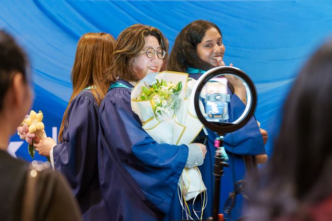 Two graduating students, one holding a bouquet of flowers, smile and pose at the 360 degree camera.