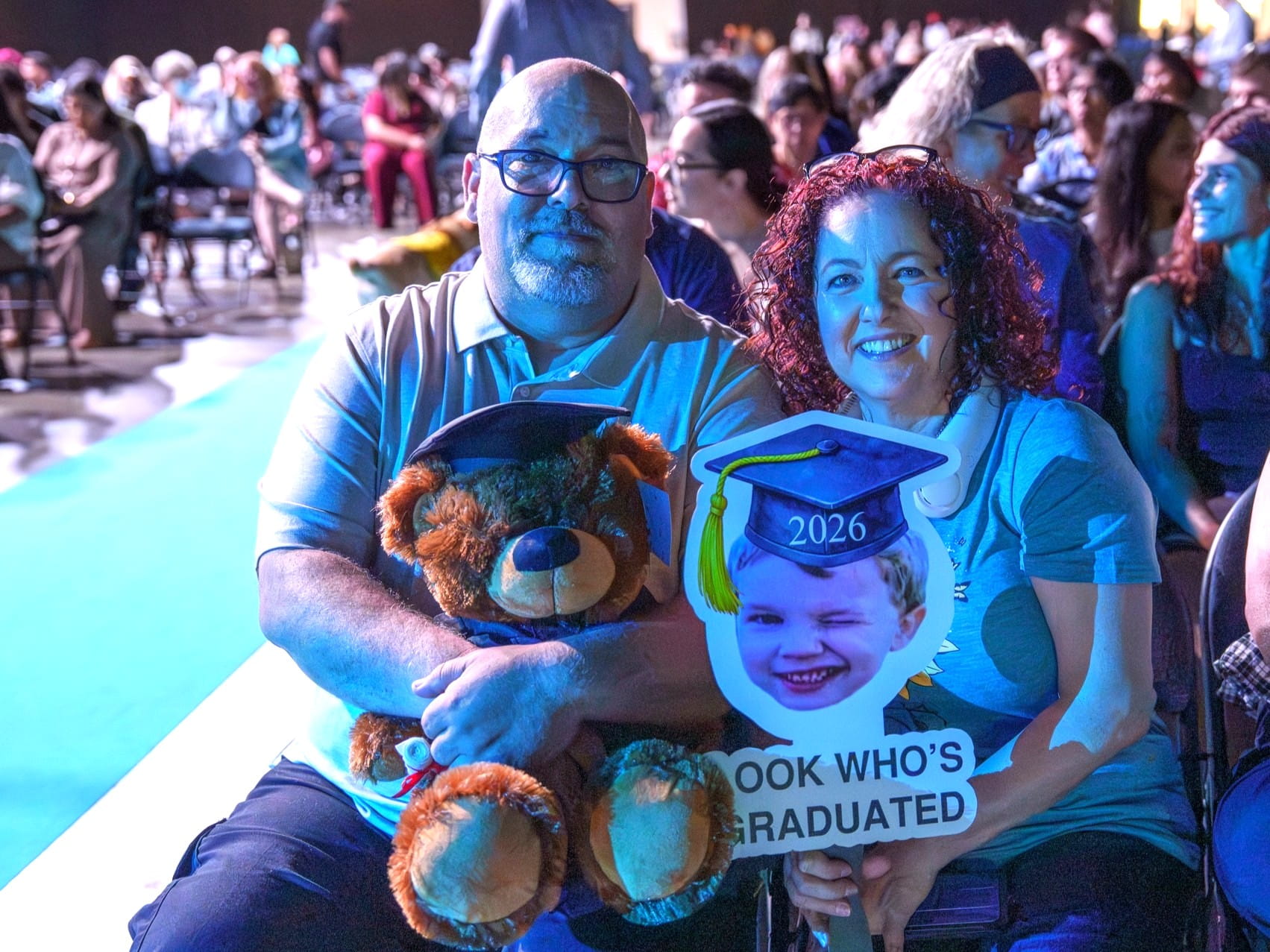 A man and woman hold a teddy bear and cardboard cutout photo of their graduating son.