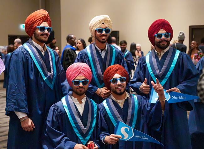 A group of graduating students, wearing robes, turbans and sunglasses, smiles at the camera.
